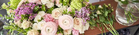 En person holder en kurv fyldt med farverige blomster, herunder roser, ranunkler og liljekonvaller. Der er også flere friske blomster på bordet, og stemningen er lys og indbydende. 